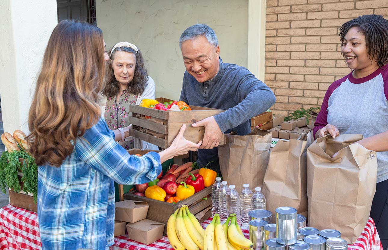 People gather around a table with fresh produce, canned goods, and paper bags at an outdoor food stand.